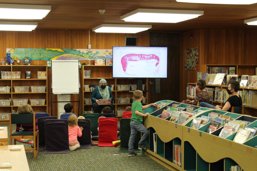 The library is located on the lower level of the Highcroft Campus in room 118. The library is a beautiful space with large, open window and plenty of space to relax and read. Ariana Hussain, the lower school librarian, is shown above reading a book to a pre-kindergarten class using an updated screen that displays the book for all students to see. Students appear intrigued and focused on the story. 